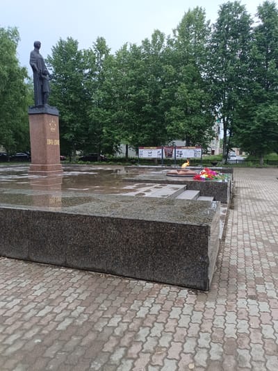 In the city of Buy, Russia, a war memorial stands at a memorial complex. The memorial features a statue of a woman holding a child, atop a granite pedestal inscribed with "1941-1945". In front of the statue, an eternal flame burns next to a bed of colorful flowers. The ground around the memorial is paved with interlocking bricks and features several shallow pools of water, suggesting recent rain. In the background, lush green trees line the perimeter of the memorial site, with buildings and parked cars visible beyond them. The sky is overcast.