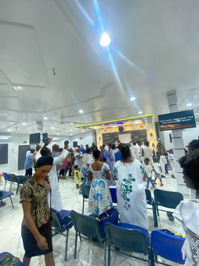 Inside a brightly lit church, people stand with raised hands, seemingly in prayer or worship. They wear a variety of clothing, including traditional white garments, floral patterns, and casual wear. Rows of dark blue plastic chairs provide seating. A small Christmas tree is visible near the altar area, where a screen displays text. The architecture features high ceilings and decorative lighting fixtures.