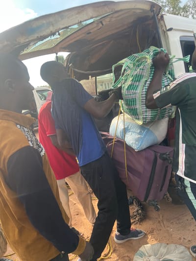 In the city of Gbaza, Nigeria, four adult males are engaged in the loading of cargo into the rear compartment of a white utility vehicle under a bright daytime sky. The scene is set on a light brown, sandy ground surface.

The rear hatch of the white vehicle is open and elevated, revealing a cargo area where several items are already stowed. A large purple wheeled suitcase lies on its side, topped by a light blue and white woven sack.

In the central foreground, a male individual wearing a dark blue short-sleeved shirt and black trousers is positioned with his back partially to the viewer, leaning into the vehicle's opening. He is actively pulling a large green and white checkered bag upwards with a yellow rope, demonstrating physical exertion. He wears black gloves on both hands. To his right, another male, partially visible and wearing a dark green shirt with white and black striped accents, assists in handling the green and white bag.

To the left of the central figure, a third male is seen from the chest down, wearing a red short-sleeved polo shirt and light-colored trousers, positioned behind the man in blue. Further to the left, a fourth male, mostly visible from the back and left side, wears a mustard yellow collared shirt over a black long-sleeved garment, observing the loading process.

A coil of yellow rope is visible on the ground near the purple suitcase. In the background, a white pickup truck or SUV is parked, and distant buildings are faintly visible under the clear sky. The interior of the vehicle's open hatch shows signs of wear and exposed components.