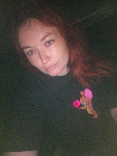 The image shows a close-up, overhead shot of a woman with reddish-brown hair. She is wearing a dark t-shirt with a cartoon image of Jerry from Tom and Jerry holding a pink heart. The lighting is dim, and the image has a grainy quality. The woman's face is partially visible, with her eyes looking towards the camera. Her expression is neutral. The background is dark and indistinct. There is no visible information in the image that would suggest a specific location in Novocherkassk, Russia.