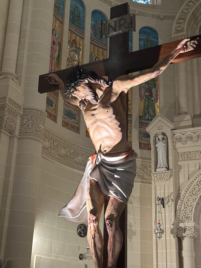 A large, three-dimensional crucifix statue is centrally positioned within the Basilica of the National Shrine of the Blessed Virgin of Ta' Pinu, Għarb, Malta. The statue depicts Jesus Christ crucified on a dark brown wooden cross, with a metallic plaque inscribed "INRI" affixed above the figure's head. The figure of Christ, rendered in realistic detail, displays a crown of thorns, visible wounds on the hands, feet, and torso (including a side wound), with simulated blood streaks prominently depicted on the limbs and body. A white loincloth is draped around the waist, and the physique appears emaciated, showing defined ribs.

The background reveals the ornate interior architecture of the church. Light-colored stone walls feature decorative pilasters with carved capitals and bases. Arched niches in the upper wall sections contain colorful mosaic or fresco panels depicting religious figures, including angels, with partial Latin inscriptions visible such as "AZANUS," "LIVA SPECIOSA," and "SAPIENTIAE." Elaborate carved friezes with floral and foliate motifs adorn the walls horizontally. A small white statue of an angelic figure with clasped hands is set within a decorative niche on a pilaster to the right of the crucifix. A silver-colored hanging lamp or censer with red accents is suspended from the wall on the right. Direct lighting illuminates the crucifix, casting shadows on the surrounding architectural elements.