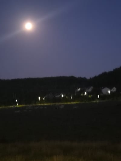 The image depicts a nighttime scene with a full moon shining brightly in a dark blue sky. A faint light streak emanates from the moon. Below the sky, a dark, tree-covered hillside is visible. In front of the hillside, a row of illuminated houses with glowing windows can be seen. Along the edge of the houses, several outdoor lights are lit, casting a yellow glow. The foreground is a dark, blurred area, possibly a field or grass, with some hints of dry grass or vegetation. The overall impression is a peaceful, rural or suburban setting at night. There are no people or animals visible in the image. The exact location in Differdange, Luxembourg, is not identifiable from the visual information alone.