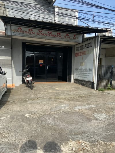 The image shows the exterior of a law firm in Purwakarta, Indonesia, located at the coordinates -6.555 latitude and 107.437 longitude. The firm is called "WARISON SIMARMATA, S.H., M.H." and is located at Jln. Jend. A. Yani No. 124/65. The sign above the entrance reads "ADVOKAT" and the sign to the right reads "KANTOR ADVOKAT/PENGACARA". A black motorcycle is parked in front of the entrance, with a sign in Indonesian that reads "BUKA" (open). The entrance has two glass doors with black frames. There is a partial view of a white car parked to the left of the entrance. The building is surrounded by a concrete sidewalk with a small patch of grass visible on the right side of the image. There are multiple electrical wires running across the top of the image. The environment is urban, with buildings and sidewalks visible in the background.