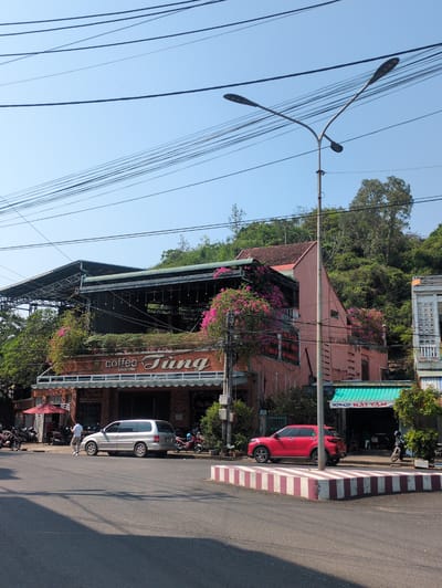 The image shows a street scene in Tuy Hòa, Vietnam. A coffee shop, "Coffee Tùng since 1980", is prominently featured, adorned with greenery and flowering vines. Next to the coffee shop is "NHÔM KÍNH HẢI TÂM" in Vietnamese text, which is a metal and glass store. Red and silver cars are parked along the curb in front of the buildings. A street lamp stands tall, with numerous power lines crisscrossing the sky above. A hillside with trees is visible in the background. A person is standing on the sidewalk by a silver car. The road appears to be recently paved.