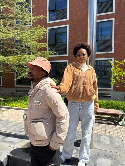 Two individuals are outdoors, likely in an urban setting, based on the presence of a multi-story brick building with windows and a silver pole that could be part of a railing. The ground appears to be paved with various materials, including gray tiling.

The first individual, standing, has dark curly hair and is wearing sunglasses, a white turtleneck, a brown furry jacket, and light blue jeans. The second individual is wearing a peach-colored bucket hat and a light beige puffer jacket. The standing person is holding the shoulder of the person wearing the hat.

The background includes greenery, suggesting a park or garden area, along with parts of a dark green bench. It's difficult to discern the specific location as Daxing District, China, without more distinctive landmarks.