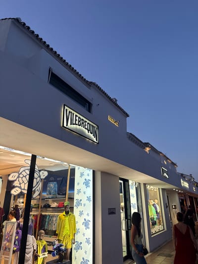 The image captures an outdoor street scene in Marbella, Spain, at dusk. The architecture is predominantly white, with a tiled roofline and illuminated storefronts. A prominent illuminated sign for "VILEBREQUIN" is visible, along with another for "HERMES" further down the facade. Store windows display mannequins dressed in brightly colored clothing, with a turtle motif featured in the Vilebrequin store's display. Several people are present, walking along the sidewalk and browsing the shops. The sky is a deep blue, indicating the late hour. The overall impression is one of a sophisticated shopping district.