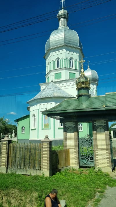 The image depicts a section of an Eastern Orthodox church in Krasnoilsk, Ukraine. The church features a prominent, tall, silver-domed tower with green accents and white architectural details. To the right of the main tower, a smaller, gilded dome with a cross is visible, part of another section of the church. In the foreground, a green-tiled roof structure covers an ornate green metal gate. This gate is flanked by brick pillars, one of which has a decorative cap. A wooden fence runs alongside the property. The ground is covered with lush green grass and vegetation. In the lower right corner of the frame, a person is seated, facing away from the camera, with their head down. Power lines are visible crossing the bright blue sky.