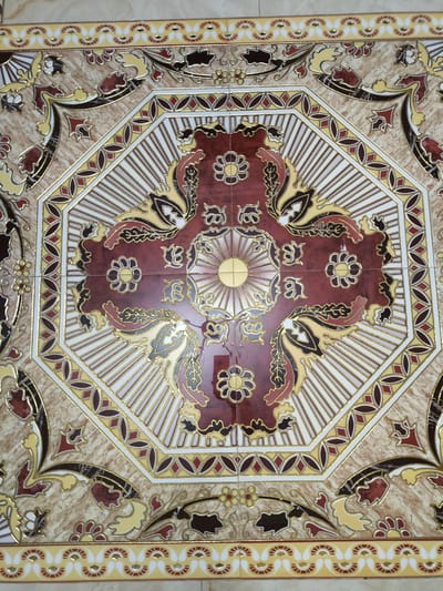 The image is a close-up, overhead shot of a decorative floor tile. The tile features an intricate octagonal design in the center, with a central motif that resembles a stylized sun or flower surrounded by radiating lines. This central design is rendered in shades of deep red, gold, and white. The octagonal border is outlined in gold and contains repeating patterns of stylized leaves and flowers in red, yellow, and dark brown, all on a marble-like, beige and brown background. The outer edges of the tile display a repeating band of alternating yellow and white shapes, accented with small red and gold details. The overall aesthetic is ornate and traditional.
There is no information available in the image to suggest a specific location in Yuzhno-Sukhokumsk, Russia. The image depicts a decorative element and does not contain any contextual clues about its placement within a city or country.