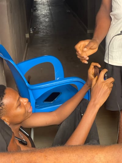 Two people are using their smartphones. One person is sitting on a blue plastic chair, with their phone tucked into the backrest. This person is wearing black pants, a black shirt, and a silver necklace. The other person is wearing white and black clothing. The scene is located in a garage, with a concrete floor and wall on the left.