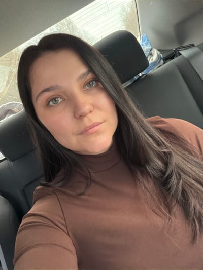 An adult female with long, dark brown hair and greenish-brown eyes is positioned in the rear passenger seat of a vehicle. She is wearing a form-fitting, solid brown, high-necked long-sleeve top and is looking directly at the camera. The car's interior features dark-colored, likely leather or synthetic, seats and headrests. A light-colored patterned fabric object is draped over the seat to the subject's left, and a blue and white patterned fabric is visible behind the headrest on the right. A seatbelt mechanism is visible on the right side. Through the rear window, bare tree branches and an overcast sky are visible, indicating an outdoor, possibly wintry or early spring environment. The specific location within the city of Vozhoi, Russia, cannot be determined from the visual information provided.
