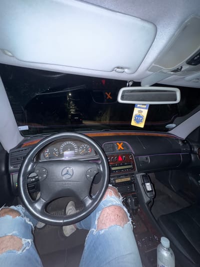 The image shows the interior of a car, likely a Mercedes-Benz, with the perspective from the driver's seat. The dashboard is visible, featuring a steering wheel with the Mercedes-Benz logo, a speedometer, and a radio. There are also decorative purple LED lights accenting the interior. A "VIP" air freshener hangs from the rearview mirror. The driver's legs, clad in ripped jeans, are visible in the foreground. The car appears to be parked or stopped at night, with the dark exterior visible through the windshield, suggesting a location possibly outside of a well-lit area. The context of Smolyan, Bulgaria is not evident from the visual information in the image.