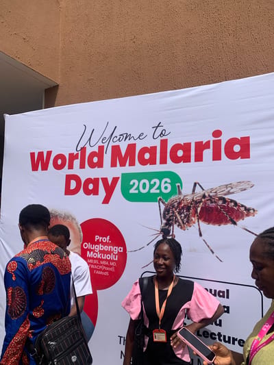 An outdoor scene captured with a slightly rotated perspective shows several individuals gathered around a large banner advertising "World Malaria Day 2026." The banner, predominantly white with red and green text, features a prominent image of a mosquito.

In the foreground, two individuals are partially visible, one wearing a vibrant blue and orange patterned top and carrying a black checkered bag, and another with their back to the camera, wearing a white t-shirt. To the right, a woman in a black and pink top smiles faintly, looking slightly upwards. She wears an ID badge around her neck, which appears to have "KWARA STATE UNIVERSITY" visible. Another person, seen from the chest up, is on the far right, holding a pink-cased smartphone. A person in the upper right corner, wearing a light green top with a pink lanyard, is also partially visible, with "University of Ilorin" discernible on the lanyard.

The banner's text includes "Welcome to World Malaria Day 2026." A red circular element on the banner introduces "Prof. Olugbenga Mokuolu," followed by his credentials: "MB;BS., MBA, MD, FWACP(Paed), FAMedS." Other partial text on the banner includes "sual," "Lecture," "CLS," "University of Ilorin," and "TH N." The background beyond the banner is a textured, light brown wall. The lighting suggests it is daytime. The people appear to be either attending or arriving at an event related to World Malaria Day, possibly in Ilorin, Nigeria, given the mention of Kwara State University and the University of Ilorin.