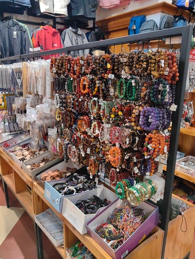 This image depicts the interior of a retail store in Sa Pa, Vietnam, primarily featuring merchandise displays. In the midground, a black metal grid stand presents hundreds of beaded bracelets in various colors, including green, purple, orange, brown, and multicolored, many individually tagged with price labels indicating Vietnamese Đồng (VND). To the left of this central display, wooden shelving units hold multiple open boxes containing miscellaneous jewelry components and small items, along with packaged necklaces and earrings suspended from hooks. The upper background shows racks of assorted outerwear, including red, white, grey, and patterned jackets, as well as blue and grey backpacks on a wooden ledge. The lower foreground includes more wooden shelving with open cardboard boxes filled with a variety of string bracelets, bangles, and other small accessories. The floor features a geometric pattern of light and dark brown tiles. No individuals are visible within the scene.