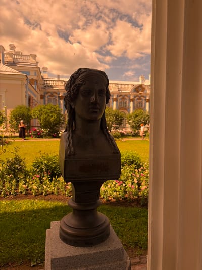 The image shows a bust of a woman with wavy hair, positioned in the foreground and facing left. The bust is made of dark material and rests on a pedestal that is partially visible. Behind the bust, a garden with lush green grass, blooming pink and yellow flowers, and trees is depicted. In the background, a grand, ornate building with blue and white architectural details and arched windows stands under a cloudy sky. The lighting suggests it might be late afternoon or early evening, with warm, golden tones cast over the scene. A person in dark clothing can be seen in the middle ground, walking through the garden. The image is oriented vertically, rotated 90 degrees counter-clockwise. The location appears to be a formal garden with classical architecture, consistent with Pushkin, Russia.