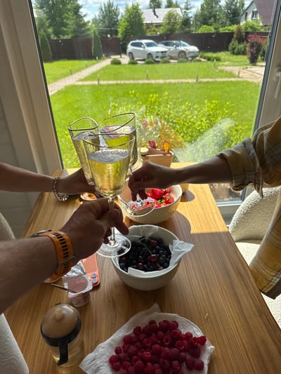 The image captures a close-up of a wooden table with several glasses of champagne being raised for a toast. In the foreground, a person's hand wearing an orange wristwatch is holding one of the glasses. To the left, another hand with a silver bracelet is also holding a glass. Between these hands, a bowl of strawberries and a bowl of blueberries and cherries are visible. There is also a small container of yogurt with a spoon, an orange smartphone, and a baby bottle with a yellow liquid. In the background, a large window overlooks a lush green yard with a pathway, trees, and a fence. Two white cars are parked in the yard, and a house is visible in the distance. The scene appears to be set in a domestic environment in Борисьево Днп, Russia, possibly during a celebration or a casual gathering.