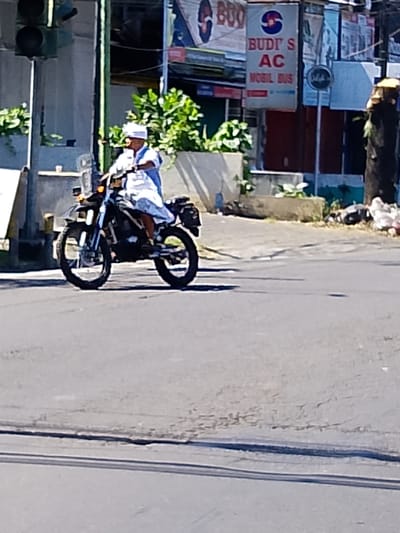 The image captures a street scene in Tabanan, Indonesia, on a bright, sunny day. A man wearing a white cap and traditional white clothing is riding a dark-colored motorcycle. The motorcycle appears to be a dirt bike or dual-sport model, with knobby tires and a high ground clearance. The man is positioned in the center of the frame, with the motorcycle moving from left to right.

In the background, there are several buildings with various signs. One prominent sign reads "BUDI'S AC" and "MOBIL BUS," suggesting a business related to air conditioning and bus services. Another sign has text that is partially obscured but appears to be in Indonesian. There is also a traffic light visible on the left side of the image. Green foliage is present along the buildings, indicating some landscaping. The street is paved, with visible lane markings. The overall environment suggests an urban or semi-urban setting within Tabanan.