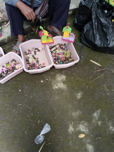 On a mossy, concrete surface in Gebang, Indonesia, a person is seated with three pink plastic containers in front of them, filled with shells and colorful beads. Two small, plastic toy houses sit atop one of the containers. A black plastic garbage bag is present nearby. The person is wearing sandals, blue pants, and a light blue long-sleeved shirt, with a brown shoulder bag beside them. A small, tied-up plastic bag and scattered leaves are on the concrete surface.