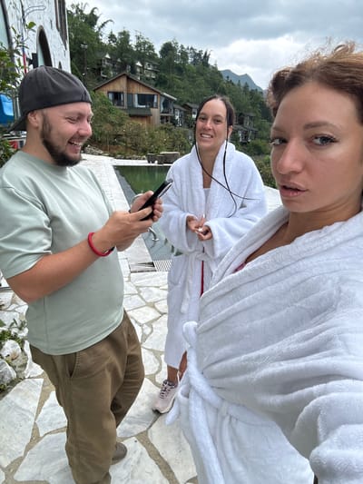The image depicts three individuals outdoors in the city of Sa Pa Ward, Vietnam. Two women are wearing white bathrobes. One woman is holding something small in her hand. A man with a beard and a cap is looking at his phone. The ground is paved with irregular, white stones. In the background are trees, houses, a pool of water, and a mountain range. The sky is overcast.