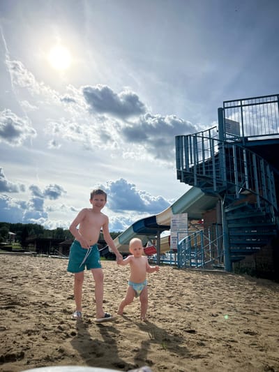 In the city of Yubileyny, Russia, two young boys are walking on a sandy beach. The older boy, wearing teal swim shorts, is holding the hand of the younger boy, who is wearing a diaper. Behind them, a blue metal spiral staircase leads up to a water slide. The sky is filled with dramatic clouds, and the sun is shining brightly. In the background, there are trees and buildings.