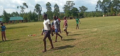The image depicts a group of children playing in a grassy field in Bungoma, Kenya, at the coordinates 0.553 latitude, 34.773 longitude.  The children are all wearing casual clothing and are walking in a line, except for one child standing off to the side, looking on. The field is surrounded by trees and a building with a green roof, possibly a school.  The children are walking in a line, and appear to be engaged in a game or activity. The day is sunny with blue sky and some puffy white clouds in the background. The field is covered in lush green grass, and the trees surrounding the field are green as well.