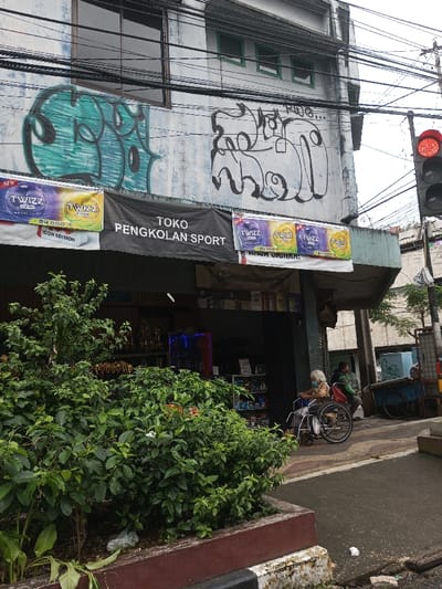 The image depicts an urban street scene featuring a two-story commercial building. Contrary to the requested location of Vaatharawattai, Sri Lanka, a prominent black banner stretched across the ground floor explicitly displays "TOKO PENGKOLAN SPORT" in Indonesian. The building's light-colored upper facade is marked with two distinct graffiti tags: a large, teal-green filled design on the left, and a black outline design with partial text "Pug..." on the right. Multiple dark utility cables are strung horizontally across the upper portion of the frame.
Beneath the main banner, smaller white banners advertise "TWIZZ" products, featuring "ROYAL LUCH" branding and a price of "24.000." The ground floor shop entrance is open, revealing shelves and a blue-lit display unit inside.
In the foreground, a rectangular planter box containing dense green foliage separates the viewer from the sidewalk. A white plastic bag is visible discarded within the foliage. The planter box features a brown top edge over a weathered white and black base.
On the sidewalk to the right, an individual with white hair is seated in a black wheelchair, facing towards the shop. This person is wearing a light-colored top and has a red bag positioned on their lap. Further back on the sidewalk, a second individual in a dark jacket is partially visible, seated near a light-blue, two-wheeled pushcart. A red traffic light, illuminated to indicate 'stop,' is visible on the far right of the frame, mounted on a pole. The street surface in the lower right appears to be dark asphalt.