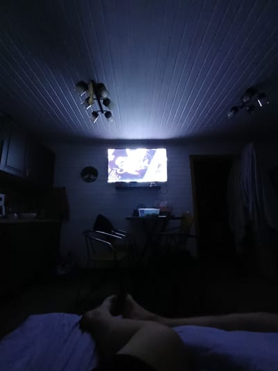 The image depicts an interior scene, possibly a living area or a bedroom, with a television on and illuminated. The lighting is dim, suggesting it is nighttime or the room is otherwise dark. The ceiling appears to be paneled. There are light fixtures visible on the ceiling. In the foreground, a person's legs are visible, lying on what appears to be a blue blanket or bedspread, indicating a seated or reclining individual. The room contains furniture, including chairs and a table, and a kitchen area with cabinets is visible on the left. A decorative circular object is mounted on the wall. The scene is a private, indoor setting. The location in the city of Koledino, Russia, is not discernible from the visual information provided in the image.
