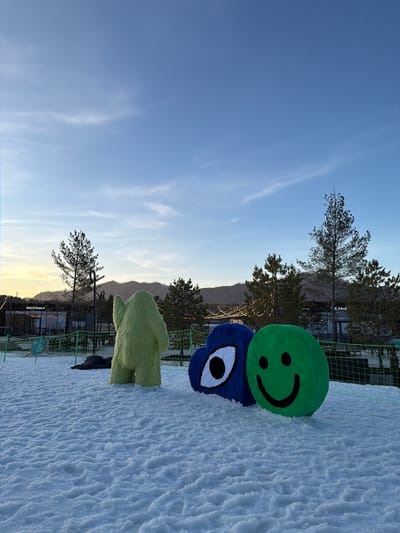 The image shows a snowy outdoor scene in Озёрное, Russia. A green smiley face and a blue monster-like plush toy are stacked on the snow, near a green fence. In the background, there are trees, a building with lights, and mountains under a blue sky.