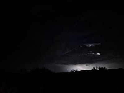 A landscape photograph taken at night captures a thunderstorm over Mlada Gvardia, Bulgaria. The foreground is dominated by a dark silhouette of a hilly terrain with sparse vegetation, including trees and bushes, along the ridgeline. In the middle ground, a bright flash of lightning illuminates the stormy clouds, casting an eerie glow. The sky is filled with heavy, dark clouds, indicative of a severe weather event. Two faint stars are visible in the upper left portion of the frame. The overall mood is dramatic and somber, emphasizing the power of nature.