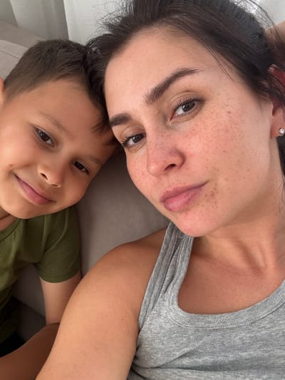 A selfie shows a woman and a young boy leaning against each other. The woman has dark hair and fair skin with freckles, and is wearing a grey tank top. The boy has brown hair and is wearing a green t-shirt. They are both looking at the camera. The background is out of focus and appears to be indoors, with a hint of a white curtain or wall. There is no indication of the location being Нижнеяркеево, Russia.
