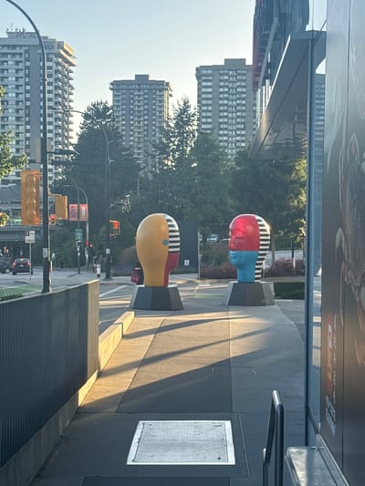 The image depicts an outdoor urban scene in Burnaby, Canada, with two large, abstract head sculptures prominently displayed on a sidewalk. The sculpture on the left is primarily yellow with red and black striped accents on one side of its face. The sculpture on the right is red on the upper half of its face and blue on the lower half, also featuring black stripes on one side. Both sculptures are mounted on dark gray, multi-sided bases.

In the background, several tall, multi-story residential buildings with numerous windows are visible, suggesting a dense urban environment. Trees with green foliage are interspersed between the buildings and around the sculptures. Traffic lights and street signs are present, indicating a public thoroughfare. One traffic light shows a yellow signal. A car is visible driving in the background on the left side of the image.

The foreground shows a paved sidewalk with a concrete curb and a dark, ribbed metal barrier on the left. The ground is a mix of concrete and asphalt, with a metal grate embedded in the pavement. Sunlight casts distinct shadows across the sidewalk, indicating the time of day is likely late afternoon or early morning. A portion of a building with a reflective glass facade is visible on the right side of the image, with a large advertisement poster partially visible. No specific individuals are clearly identifiable as the primary subjects, though several blurry figures can be seen in the mid-ground, suggesting the presence of pedestrians.