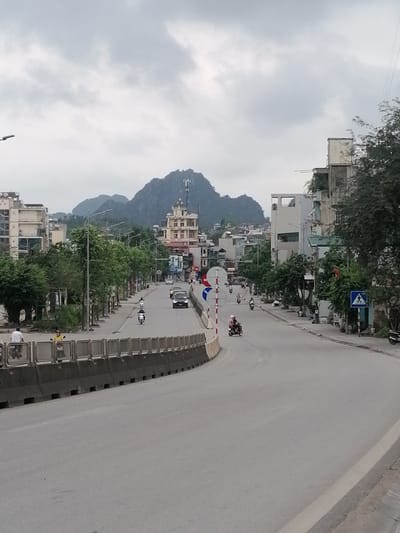 The image shows a street scene in Ha Tu Ward, Vietnam. A mountain with a telecommunications tower on top rises in the background. Buildings line both sides of a curved road, with trees interspersed. Several people are visible; some are on scooters and others are walking along the side of the road. A gray concrete barrier runs along the left side of the road. Traffic includes a black car and various scooters. The sky is overcast with gray clouds. A pedestrian crossing sign is seen on the right side of the road.