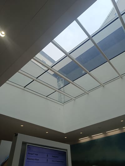 The image shows an interior view looking upwards towards a large, modern glass atrium ceiling. The ceiling is supported by a white structural frame and consists of large glass panels. Sunlight filters through the glass, illuminating the space. Below the atrium, a digital display board is visible, showing a schedule with names like "George F Jupp" and "Rosemary Hilson," along with room numbers like "Imaging Door" and "Clinic C Treatment Room." This suggests the location is likely a hospital or clinic, possibly within the city of Cambridge, United Kingdom, given the general aesthetic and information displayed. The walls are painted a neutral color, and there are recessed lights and ceiling fixtures visible. In the background, through the glass, parts of a building with windows and a striped facade can be seen. A metal structure, possibly a ladder or scaffolding, is also visible through the atrium.