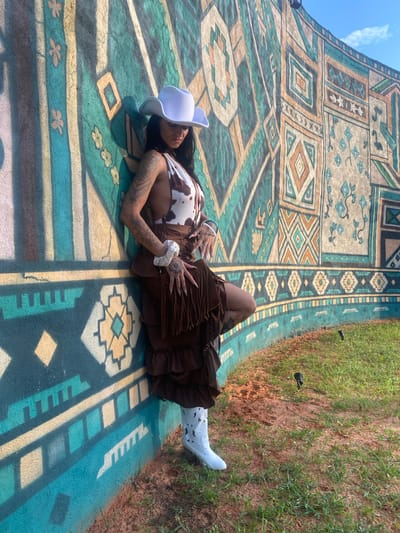 A woman in a cow-print bikini top, brown fringe skirt, and white cowboy boots leans against a vibrantly painted mural. The mural features a large, colorful, geometric pattern with turquoise, yellow, and orange hues. The woman has long, dark hair and numerous tattoos on her arms. She is wearing a white cowboy hat and her hands are positioned near her skirt. The ground in front of the mural is covered in a mix of reddish-brown soil and green grass, with a few small black spotlights visible in the grass. The scene appears to be outdoors under a bright sky. The location is the Mui Ne Sand Dunes in Mui Ne, Vietnam, specifically a roadside attraction featuring this mural.