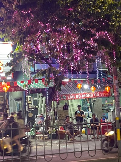 The image depicts a nighttime scene on a street in Thuận An, Vietnam, likely a commercial area given the presence of a food establishment. A tree adorned with numerous pink and white string lights hangs over the sidewalk, creating a festive ambiance. Below the tree, a storefront with a striped awning displays a sign that reads "LẨU BÒ MÔNG CỔ 199K," indicating a beef hot pot restaurant with a price of 199,000 Vietnamese Dong. Several people are visible outside the establishment. Three men are seated on low stools, appearing to be customers. One man stands and holds a large jar, possibly serving food. In the foreground, a metal fence separates the street from the sidewalk. A scooter is parked nearby, its headlight illuminated. The overall impression is of a lively evening scene in a Vietnamese city.