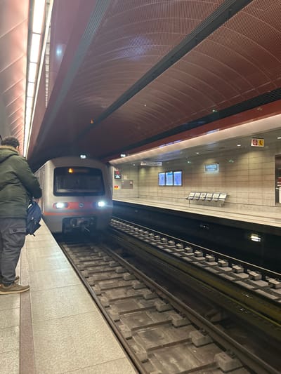 The image displays an underground subway station platform and tracks, photographed from a high angle looking down the length of the station. The photograph itself is rotated 90 degrees counter-clockwise.
In the immediate foreground, a person is lying prone on the light grey tiled platform. They are wearing a dark green padded jacket, dark grey cargo pants, and light brown shoes. A dark blue backpack rests near their head.
A modern subway train, predominantly white and grey with an orange stripe, is stopped at the platform. Its bright front headlights are illuminated. The train's front portion is visible, facing towards the right side of the frame.
To the left, a distinctive architectural feature of the station is a curved, reddish-brown wall or ceiling with a textured, perforated surface. This structure incorporates long, linear light fixtures. The subway tracks, complete with sleepers and ballast, occupy the right side of the image.
Across the tracks, on the opposing platform, several station amenities are visible, including light-colored benches and multiple digital information screens. Various signs are mounted on the station walls; one prominent sign clearly displays "ΚΟΡΥΔΑΛΛΟΣ" (Koridallos) in Greek, indicating the station's name. Below this, partially visible Greek text likely indicates a direction, such as "προς Πειραιά" (to Piraeus). Other emergency signs, including a red stop button and a yellow telephone symbol, are also present. The station is illuminated by artificial lighting, suggesting it is indoors and the external time of day is not a factor.
