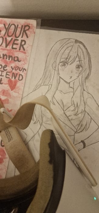 The image depicts an indoor scene featuring several objects arranged on a flat, light-colored surface. A hand-drawn pencil sketch of an anime-style female character occupies the right side, rendered from the chest up with long hair, large eyes, and a simple V-neck garment. The drawing includes subtle details such as blushes on the cheeks and a bracelet on the left wrist. To the left of the drawing, a rectangular piece of paper or card is visible, displaying bold, partially obscured text including "YOUR," "OVER," "anna," and "be your FRIEND," accompanied by a red heart symbol, all against a mottled red background. A tan-colored, textured strap or belt, possibly made of fabric, lies diagonally across the lower-middle section, partly obscuring the drawing and the note. A small white label with fine print is visible on the strap's left end. The upper portion of the background consists of a dark, indistinct surface, likely a desk or shelf, beneath a lighter, textured wall. No people or active interactions are present. The specific location within the city of Blaye, France, cannot be determined from the visual information provided.