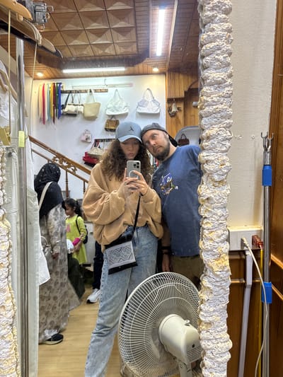 The image shows a reflection of a man and a woman in a mirror. The woman is holding a phone and taking a picture of themselves. The man is standing next to her, with his head close to hers. The reflection shows the interior of what appears to be a retail store with clothing and bags on display. Other people and items are reflected in the mirror as well. The location is in the city of Sa Pa Ward, Vietnam.