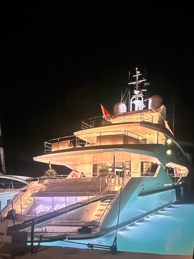 A large, illuminated yacht named "Aqua Blu" is docked at night in the waters of Municipal Unit of Lefkada, Greece. The yacht has multiple levels with interior lights creating a warm glow. The name "Aqua Blu" is prominently displayed in illuminated script on the stern, along with "VALLETTA" below it. There are railings and stairs visible on the yacht, and it is surrounded by calm, glowing blue water. The sky is dark and featureless.