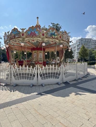 A carousel stands in a city square on a sunny day. The carousel is brightly decorated with ornate details, including painted panels depicting architectural scenes, figures of cherubs, and numerous lights. The carousel features several horses and other decorative elements. A white, decorative fence surrounds the base of the carousel. The ground is paved with light-colored square tiles, with a dark gray strip running through it. In the background, there are trees, modern apartment buildings, and a bird flying in the clear blue sky. The scene is set in Zelenograd, Russia.