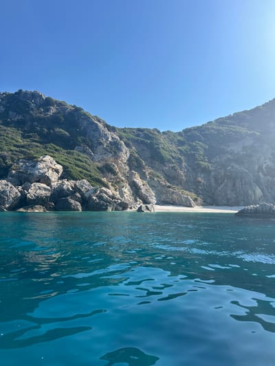 The image depicts a serene coastal scene with clear turquoise waters in the foreground. In the background, rugged, green-covered cliffs rise from the sea, revealing a small, pebble beach nestled between them. The sky above is a vibrant, cloudless blue. The perspective suggests the photo was taken from a boat or a low vantage point on the water, looking towards the shore. The scene evokes a sense of natural beauty and tranquility. The specific location within "Unknown, Greece" is not identifiable from the visual information provided.