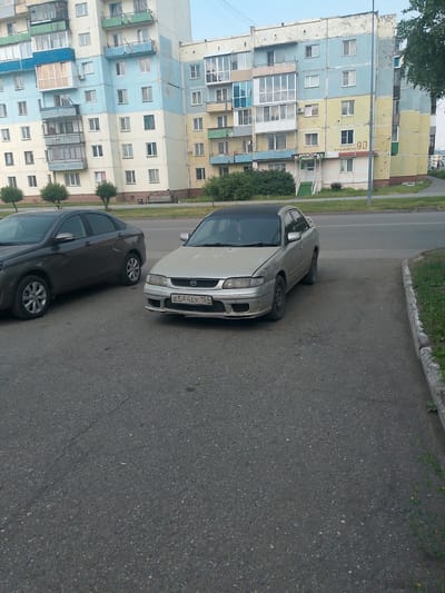 The image shows a street scene in Polysayevo, Russia, likely in the late afternoon given the soft lighting and long shadows. Two cars are parked on the asphalt. The car in the foreground is a silver sedan with a black roof and a Russian license plate reading "E544EK 154 RUS". It appears to be a Mazda. To its left is a dark gray sedan, possibly a Lada, seen from the side.

Behind the cars is a multi-story apartment building with a distinctive color scheme of light blue, cream, and yellow. The building has numerous balconies, some enclosed with glass, and visible windows. There are small trees and bushes along the sidewalk in front of the building, and a paved path leading towards an entrance.

Above the entrance of the building, some signage is visible. One sign reads "ДРУГ" (Drug), which translates to "Friend" in Russian. Another sign, to the right, has a red number "90" on it, possibly a building number or business identifier. The overall impression is a residential urban environment. The weather appears clear and calm. There are no people clearly visible in the image, and the focus is on the cars and the apartment building.