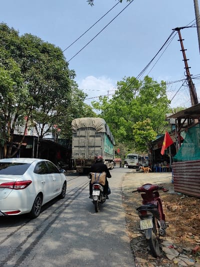 The scene depicts a street in Bình Lư, Vietnam, under a clear blue sky with some scattered clouds. The asphalt road, showing signs of wear and multiple dark tire marks, extends into the background.
In the immediate foreground on the right, a maroon scooter with a license plate "25-B1 675.66" is parked on a dirt and debris-strewn shoulder. Further along the road, a person wearing dark clothing and a red-striped helmet is riding a black scooter away from the camera. A large, light-brown package is secured to the rear of this scooter, which bears the license plate "19-H1 076.11".
To the left of the scooter, a white sedan, possibly a Toyota Vios, is either parked or moving slowly in the same direction, with its rear section visible. Ahead of these vehicles, a large semi-trailer truck, featuring a gray ribbed container covered by a light-colored tarp, occupies the center of the road and is also moving away from the viewer.
On the left side of the road, dense green trees and foliage obscure the lower sections of what appear to be residential buildings with corrugated metal roofs. On the right side, a rustic structure with a corrugated metal facade and blue tarps is visible, adorned with a Vietnamese flag (red field, yellow star) hanging from a wooden pole. Small tables and chairs are set up outside this structure. A tall, dark utility pole with several cross arms and numerous overhead wires is prominent on this side, with more wires crisscrossing the sky. Further down the road on the right, near the semi-trailer, a white minivan and a brown dog are discernible. Bright daylight illuminates the scene, casting shadows on the road.