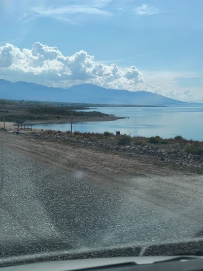 The image depicts a view from a vehicle, likely a car, looking towards a lake in Kiçi-Jargılçak, Kyrgyzstan. The foreground shows an asphalt road with worn patches of dirt. A rocky shoreline with sparse vegetation separates the road from the calm, light blue water of the lake. In the middle distance, a small beach area includes a white and red striped post and a covered structure with tables and benches. The background features a line of hazy mountains beneath a sky with scattered white clouds. Electrical poles are visible along the shoreline.