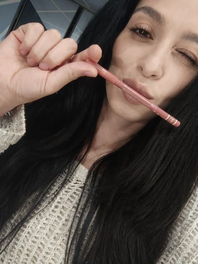 A close-up, eye-level shot captures a woman in what appears to be an indoor setting in Simferopol, Ukraine. She has long, dark hair flowing over her shoulders and is wearing a light-colored, textured knit sweater. The woman holds a pink lip liner pencil between her right hand's fingers and her slightly pursed lips, making a playful expression. She winks with her right eye while her left eye is open and looking towards the camera. She appears to have makeup on, including defined eyebrows, eyeliner, and mascara, and is wearing a gold-toned ring on her right ring finger. The background is blurred, showing indistinct architectural elements or walls. On the lip liner, the word "Creme" is visible, along with other unreadable markings and text.