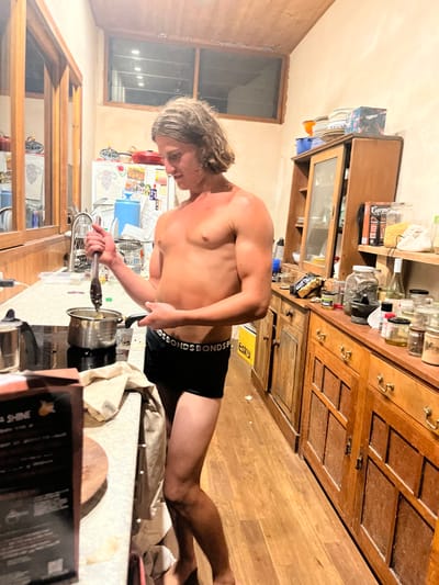 A young adult male, bare-chested and wearing black Bonds brand boxer briefs, stands barefoot in a kitchen in Stanwell Park, Australia. He has long, wavy, light brown hair and a muscular physique with visible tan lines and some redness on his shoulders. He is positioned in the center of the frame, holding a dark cooking utensil (possibly tongs) in his right hand, stirring the contents of a small silver pot that rests on a black induction stovetop. His left hand is near the handle of a dark frying pan.

The kitchen features light-colored wooden floorboards and a light-colored, possibly speckled countertop. To the left, a dark kettle or similar appliance is beside the stovetop. Further left, a wooden framed structure, possibly a window or partition, reflects internal lighting, and a white refrigerator adorned with magnets is partially visible behind it. Small horizontal windows are near the ceiling.

To the right of the male, a rustic wooden countertop and matching cabinetry extend along the wall. This area is cluttered with various items, including multiple glass jars and bottles containing spices, oils, and other foodstuffs. A dark mortar and pestle, a white bottle, and a package labeled "Carrot" are visible. An upper wooden cabinet with a glass door displays stacked dishes. A yellow "Esky" branded cooler is partially visible on the floor beneath the cabinetry. The kitchen is illuminated by artificial indoor lighting, creating a warm ambiance.