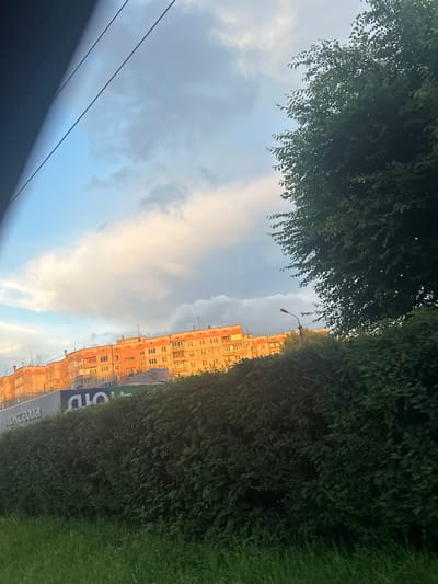 This is a rotated photograph taken from a low angle, likely from within a vehicle, looking up and outwards. The scene is dominated by a building with many windows, appearing to be an apartment block, rendered in shades of orange and yellow. The building is situated along a street lined with dense green bushes and grass. Above the building, the sky is a mix of blue and grey clouds, with some patches illuminated by the warm glow of sunset. The time of day is late afternoon or early evening, as suggested by the golden light on the building and the clouds.
In the foreground, obscuring the lower left of the image, is a dark, out-of-focus area that appears to be the interior of a car or a similar enclosed space. Two dark, diagonal lines, possibly power lines or cables, stretch across the sky. A street lamp is visible above the building, suggesting an urban or suburban setting. The overall mood is somewhat tranquil, with the soft light and the contrast between the natural elements (trees, grass, sky) and the man-made structure.
There is visible text on the side of the building, appearing to be in Cyrillic script. Part of the text reads "ПОЧТА РОССИИ" which translates to "Russian Post". The image was taken in Magnitogorsk, Russia.