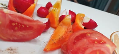 A close-up shot captures a collection of chopped vegetables on a white plate. The vegetables include slices of ripe red tomato, bright orange and yellow bell peppers, and small red radishes. The vegetables are cut into irregular shapes and are scattered across the plate, some appearing to be on skewers. A light dusting of black pepper is visible on some of the bell pepper pieces. The overall presentation suggests a prepared salad or a garnish for a dish. The background is out of focus, but hints of a patterned edge on the plate and a dark surface below suggest an indoor setting. The lighting is bright and appears to be from an overhead source, creating some shine on the moist surfaces of the tomatoes and peppers. There are no people or animals visible in the image. The image is cropped closely to the vegetables, emphasizing their textures and vibrant colors.