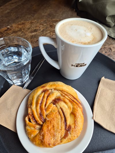 The image displays an overhead view of a table setting on a dark grey, textured tray. A white ceramic mug with the black "CAFFÈ NERO" logo contains a light brown beverage topped with white foam, positioned on the right side of the tray. To its left, a clear, faceted glass holds clear water. A slender, silver-colored utensil is partially visible between the glass and the mug. A round white plate at the bottom of the tray holds a golden-brown, twisted pastry, exhibiting a visible cinnamon swirl and sprinkled sugar crystals. Two folded, light brown paper napkins are situated on the tray: one to the left of the plate and another to the right of the mug. The tray rests on a mottled brown and beige surface, resembling marble. A dark olive-green fabric object is visible in the upper right background. The location is identified as a Caffè Nero establishment, a chain that operates within the West Suffolk district of the United Kingdom.