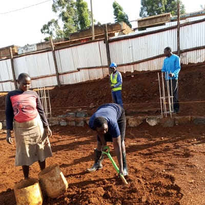 The image shows four individuals engaged in construction work in Borabu Chitago ward, Kenya. A woman in a red and black shirt and beige skirt stands near two yellow buckets. Another person is using a green shovel to dig into the reddish-brown soil. In the background, a man in a blue hard hat and a yellow reflective vest stands with a metal bar. Another man in a blue jacket stands next to reinforced steel bars. Behind them, there is a corrugated metal fence.