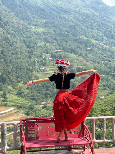 The image, rotated 90 degrees clockwise, captures a young woman in traditional attire posing on a decorative red bench or swing, set against a breathtaking backdrop of lush green mountains in Thông Nguyên Commune, Vietnam.

The woman is barefoot, standing on the red structure, and her long, pleated red skirt is dramatically fanned out and held up by her right arm. She wears a black short-sleeved top and a distinctive, elaborate red and white traditional hat adorned with intricate silver-colored motifs and small tassels. Her hair is styled in a long braid. Her pose suggests movement, perhaps dancing or simply reveling in the expansive view.

The background is dominated by vast, verdant mountain ranges covered in dense forest foliage, extending into the misty distance under a bright, sunny sky. Patches of terraced fields can be seen on the mountain slopes, indicating human activity within the natural landscape.

The red bench or swing features ornate metalwork with cut-out designs, including a circular motif and stylized text. Partially visible Vietnamese text on the structure includes "NỘI LƯU CÚ NÊN V..." and a phone number "SĐT: 09483499...". The bench is supported by what appears to be a grey concrete or stone railing structure. The scene evokes a sense of cultural celebration and scenic beauty at a mountain viewpoint.